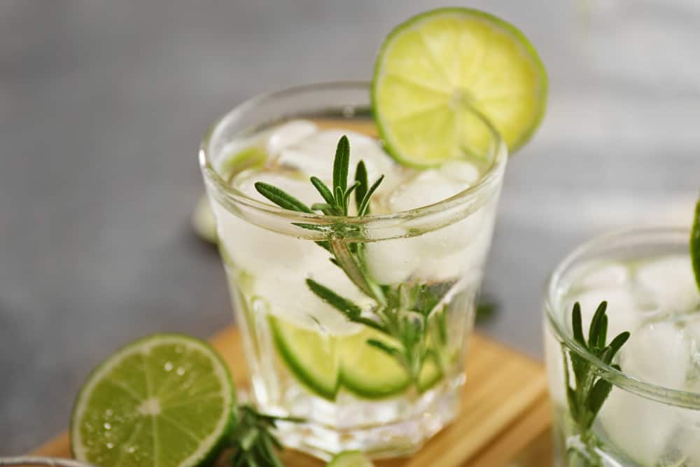 gin and tonic with lime and rosemary garnish on a wooden board surrounded by freshly cut limes