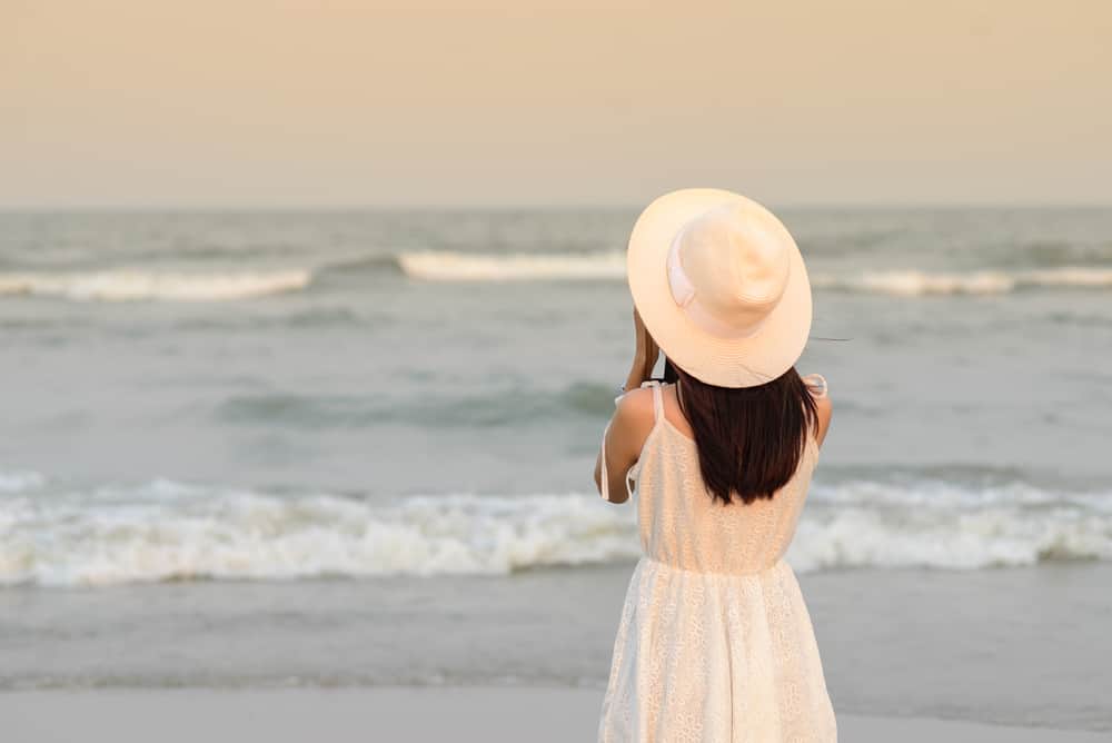 Back of woman wearing hat with white dress taking photo on the beach.